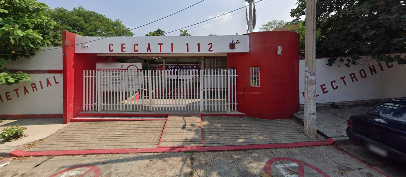 Fotografía en la que se muestra la entrada del plantel, conformada por una reja de color blanco. A los lados de la reja hay muros blancos y encima de ella hay un techado de color blanco que en letras grandes rojas dice CECATI 112. Frente a la reja hay una rampa de acceso. 